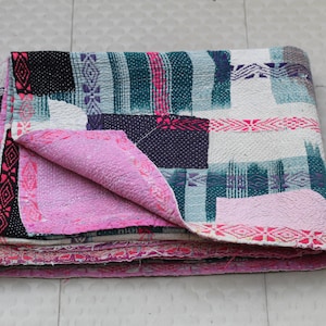 Puede incluir: Colcha de patchwork plegada con la parte inferior rosa. La colcha presenta una variedad de colores que incluyen negro, blanco, verde azulado y morado. Los bordes están adornados con un borde rosa estampado. La colcha está doblada cuidadosamente.
