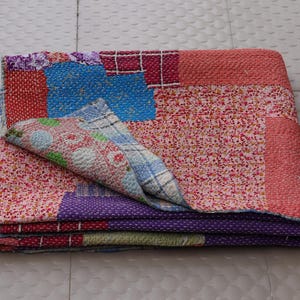 May include: A folded patchwork quilt featuring a variety of colorful fabric squares with floral, plaid, and polka dot patterns. The textile displays intricate white running stitch embroidery throughout, with visible sections of red, blue, and purple fabric.