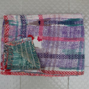 May include: A colorful, handmade textile with a patchwork design. The quilt features a mix of purple, pink, green, and white hues, with intricate stitching and a decorative border. The textile is folded, showcasing its detailed craftsmanship and vibrant colors.
