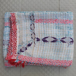 May include: A folded textile with a light blue and beige background. It has a red and white patterned border with fringe, and decorative purple and pink embroidered details. The design is geometric and textured. The textile is likely hand-woven.
