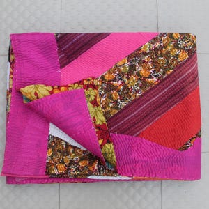 Puede incluir: Un edredón de patchwork plegado con una vibrante mezcla de colores y patrones. El edredón presenta secciones de rosa brillante, rojo y burdeos, junto con diseños florales y a rayas. Los bordes están acabados con un borde rosa liso.