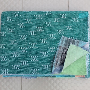 May include: A folded teal blanket with a white zigzag pattern. The blanket has a section folded back, revealing layers of fabric in shades of blue, green, and gray. The blanket is on a light gray surface.