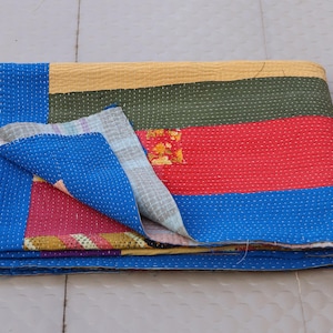 May include: A folded rectangular quilt featuring horizontal bands of yellow, dark green, and red fabric. The quilt is bordered by a bright blue fabric with white running stitch details throughout. A corner is folded back to reveal a blue patterned lining.