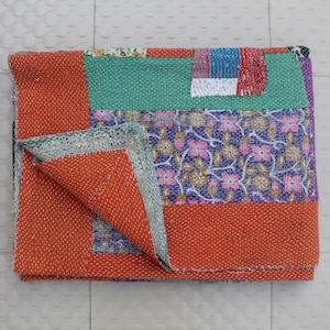 Puede incluir: Un edredón de patchwork plegado con una vibrante mezcla de colores y patrones. El edredón presenta un gran borde naranja, una sección turquesa y un centro con estampado floral en morado y rosa. Los bordes están cosidos con hilo blanco.