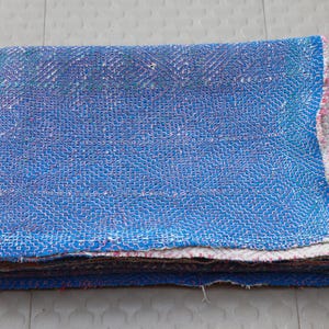 May include: A folded rectangular textile featuring a vibrant blue base color with intricate, dense white stitching patterns across the surface. The edges reveal layers of multi-colored fabric scraps, showcasing a textured, layered construction.
