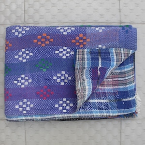 Op de afbeelding: Een opgevouwen, blauw-paarse quilt met een geometrisch patroon van witte, oranje, groene en rode vormen. De quilt heeft een contrasterende blauwe en geruite onderkant en is waarschijnlijk handgemaakt.