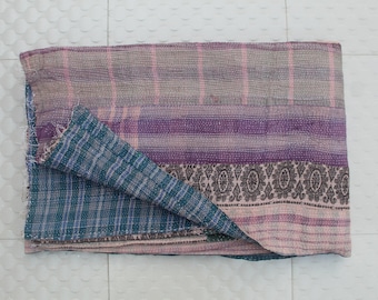May include: A folded, reversible textile with a patchwork design. The top side features stripes in shades of pink, purple, and gray, while the underside displays a teal plaid pattern. A decorative border with an ornate design is visible.
