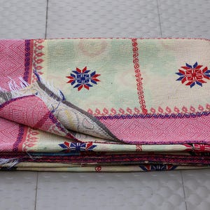 May include: A folded textile blanket featuring a cream-colored base with a prominent pink border. The fabric displays a repeating pattern of red and blue star-like motifs, vertical red lines, and visible white fringe along the folded edge.