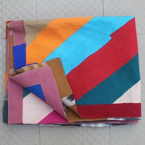 May include: A folded, multi-colored quilt with a patchwork design. The quilt features diagonal stripes in shades of orange, blue, red, teal, and pink. The edges are finished with a dark red border. The quilt is made of textured fabric.