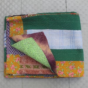 May include: A folded patchwork quilt with a variety of colours and patterns. The quilt features sections of green, blue, yellow with pink floral designs, and purple. The quilt is made of multiple fabrics.