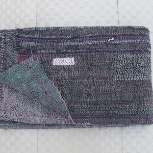May include: A folded, rectangular textile with a patchwork design. The main color is a dark gray with purple and red accents. A section is folded over, revealing a green and purple patterned underside. The textile has a textured appearance.