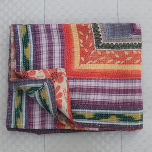Puede incluir: Un edredón plegado con diseño de patchwork en colores vibrantes. Presenta rayas moradas, naranjas y verdes, con patrones florales y geométricos. Los bordes están cosidos con hilo blanco, y se ve una esquina estampada.