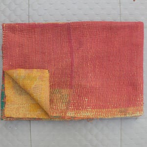 Może przedstawiać: Złożony, prostokątny materiał tekstylny o dominującym koralowo-czerwonym odcieniu. Tkanina ma teksturowany, przeszywany wygląd, ze złożonym rogiem odsłaniającym żółto-zielony wzór na spodzie. Prawdopodobnie jest to koc lub narzuta.