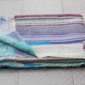 Könnte beinhalten: Ein gefaltetes rechteckiges Textil aus einem Patchwork aus mehrfarbigen Stoffschichten. Die Oberfläche zeigt horizontale Streifen in Violett-, Blau-, Grün- und Cremetönen mit sichtbaren Nähten, die sich durch das gesamte Material ziehen.