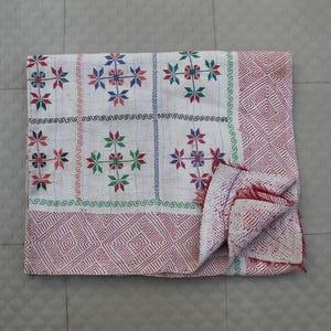 Könnte beinhalten: Weiße Steppdecke mit einem sich wiederholenden Blumenmuster in Rot, Grün und Blau. Die Steppdecke hat einen geometrischen Rand in Rot. Das Blumenmuster ist sternförmig. Die Steppdecke ist gefaltet.