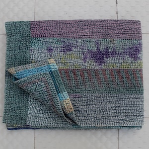 May include: A folded, multi-colored quilt with a textured surface. The quilt features horizontal stripes in shades of teal, purple, and gray, with a section folded over to reveal a blue and teal striped edge. The quilt is made of a soft material.