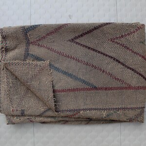 May include: Folded beige blanket with a geometric pattern of red and blue stripes. The blanket has a textured appearance and a folded corner, showcasing the stitching and fabric detail. A cozy home decor item.