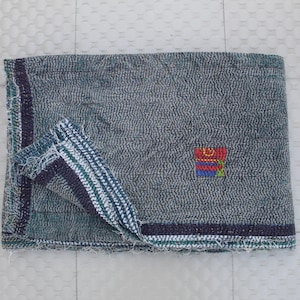 Puede incluir: Textil gris doblado con una superficie texturizada y un parche bordado colorido. Los bordes están adornados con rayas azul oscuro y verde. La esquina doblada revela un patrón de rayas en blanco, azul y verde.