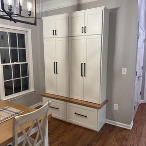 May include: White cabinet with two doors and two drawers. The cabinet has black hardware and a wood top. The cabinet is in a room with wood floors and a window.