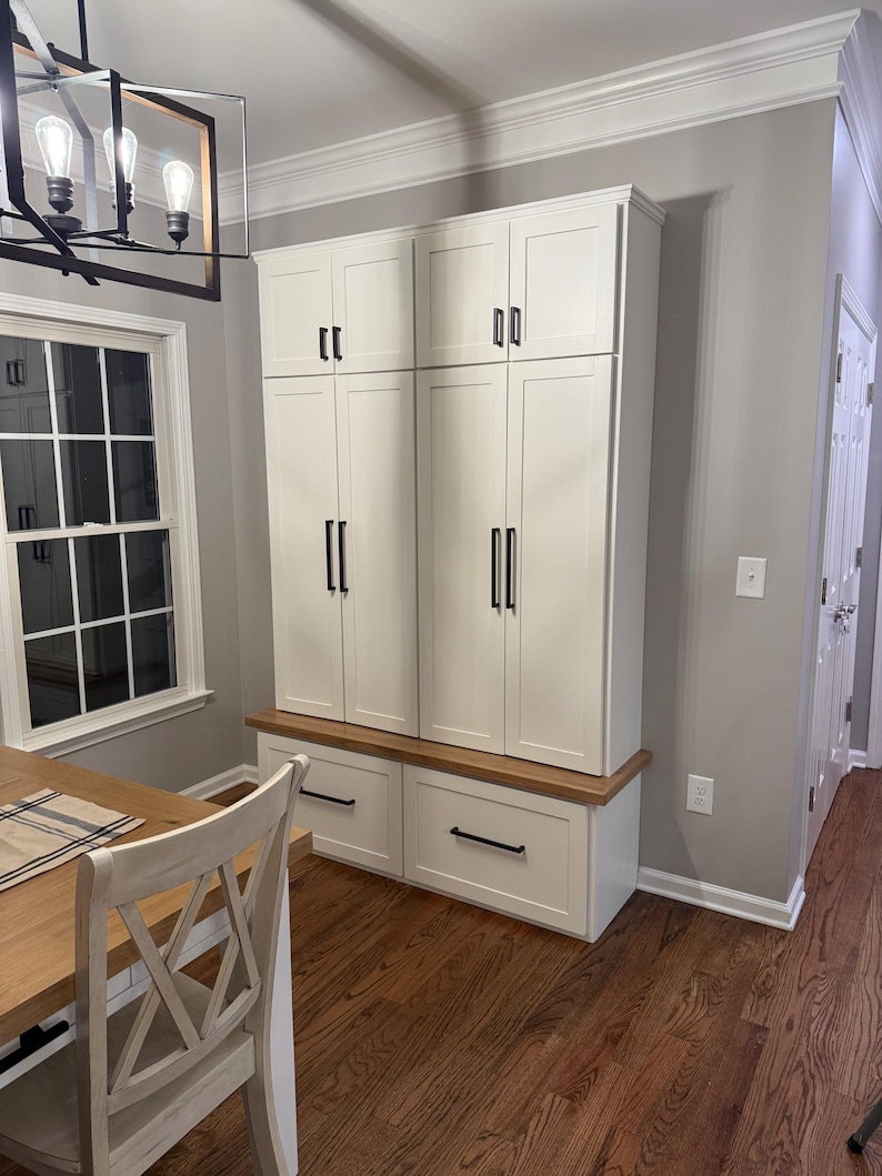 May include: White cabinet with two doors and two drawers. The cabinet has black hardware and a wood top. The cabinet is in a room with wood floors and a window.