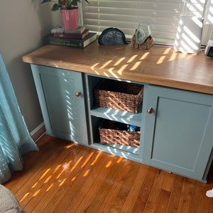 May include: A light blue cabinet with two doors and two shelves. The cabinet has a wood top and brass knobs. The shelves are filled with wicker baskets.