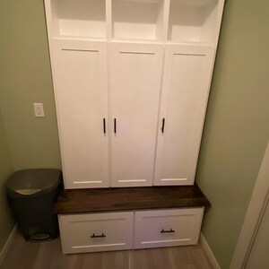 May include: White wooden storage cabinet with three doors and three open shelves above. The cabinet has a dark wood top and two drawers below. The cabinet is in a room with green walls and a gray trash can.