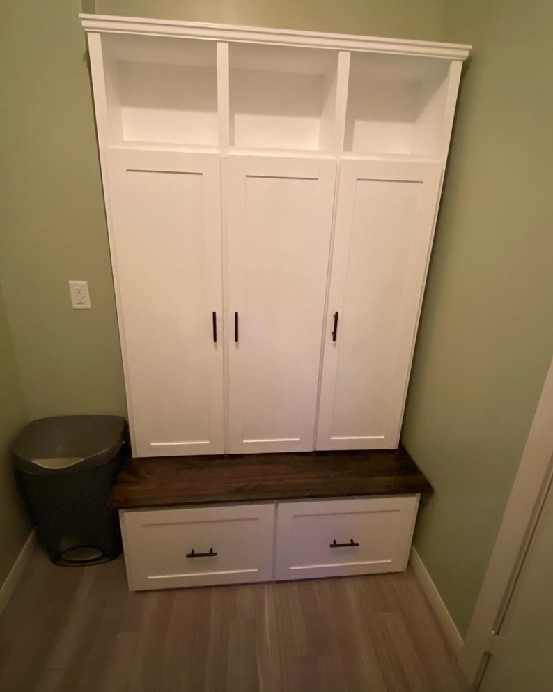 May include: White wooden storage cabinet with three doors and three open shelves above. The cabinet has a dark wood top and two drawers below. The cabinet is in a room with green walls and a gray trash can.