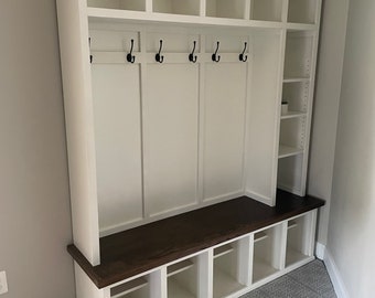 May include: A white wooden entryway organizer with a dark brown bench and cubby storage. It features six black hooks, open shelves, and cubbies for storage. The unit is designed for a home's entryway or mudroom.