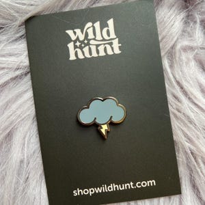 May include: A Wild Hunt enamel pin featuring a light blue cloud with a gold outline and a yellow lightning bolt. The pin is on a black card with the text "Wild Hunt" and "shopwildhunt.com" in white.