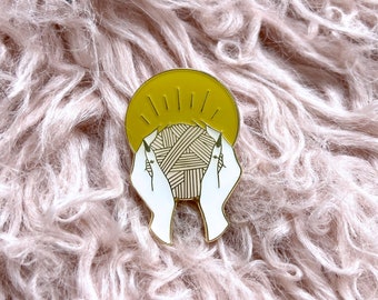 Op de afbeelding: Gouden emaille pin met een paar handen die een bol garen vasthouden met een gele zonneschijn erachter.