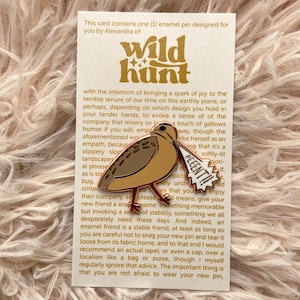 Può includere: Uno spillo in smalto dorato con un uccello marrone e bianco con un fumetto che dice "PEEENT!!" Lo spillo è su una carta bianca con testo dorato che dice "wild hunt" e una descrizione dello spillo.