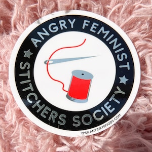 May include: A round sticker with the text "ANGRY FEMINIST STITCHERS SOCIETY" in a black and white design. The sticker features a needle and thread, and a red spool of thread. The background is a soft, pink, fuzzy texture.