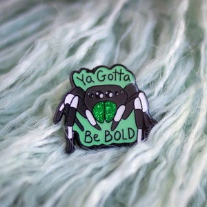 Puede incluir: Un pin de esmalte negro y verde con una araña de dibujos animados con el texto "Ya Gotta Be Bold" sobre un fondo verde.