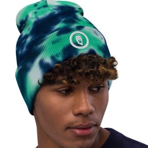 May include: A green and blue tie-dye beanie with a white circle logo featuring a stylized face. The beanie is made of a soft, comfortable material and is perfect for keeping your head warm in the winter.