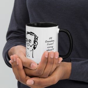 May include: White coffee mug with a black rim and handle. The mug features a black and white illustration of a person wearing headphones and the text "OK Grandma, I won't worry about it anymore."