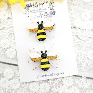 May include: Two bee-themed hair clips with gold glitter and black and yellow stripes. The clips are on a white card with the text "Girls in BloomZ Handmade Hair Accessories" and "GirlsinBloomZ.etsy.com".