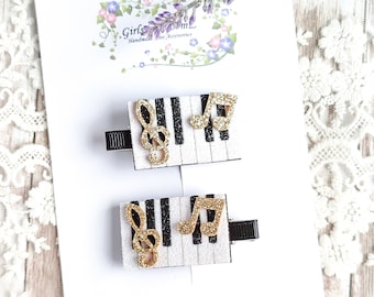 Puede incluir: Dos pinzas para el pelo de teclas de piano en blanco y negro con notas musicales doradas con purpurina. Las pinzas están en una tarjeta blanca con el texto "Girls in Bloom Handmade Hair Accessories" y "GirlsinBloomZ.etsy.com".