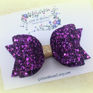 May include: A purple glitter hair bow with a gold center. The bow is on a white card with the text "Girls in BloomZ Handmade Hair Accessories GirlsinBloomZ.etsy.com".
