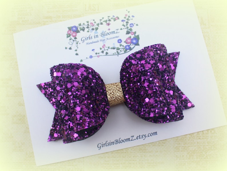 May include: A purple glitter hair bow with a gold center. The bow is on a white card with the text "Girls in BloomZ Handmade Hair Accessories GirlsinBloomZ.etsy.com".