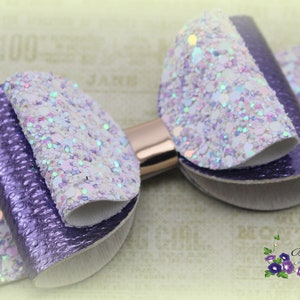 May include: A handmade hair bow featuring a layered design with purple and white glitter accents. The bow has a metallic gold center and is suitable for various occasions. The bow is from Girls In Bloom Z.