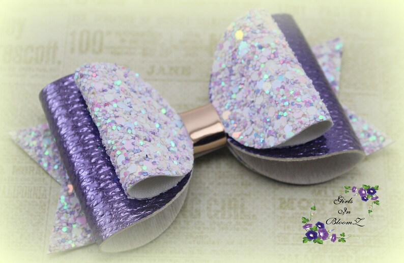 May include: A handmade hair bow featuring a layered design with purple and white glitter accents. The bow has a metallic gold center and is suitable for various occasions. The bow is from Girls In Bloom Z.
