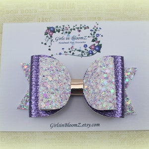 May include: A handmade hair bow featuring a glittery white and purple design. The bow is attached to a gold-colored barrette. The bow is on a white card with the text "Girls in BloomZ" and a floral design.