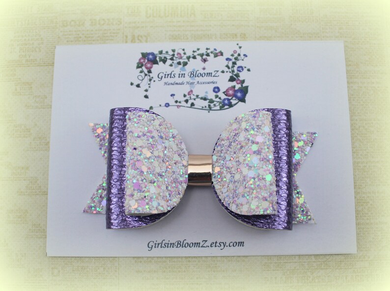 May include: A handmade hair bow featuring a glittery white and purple design. The bow is attached to a gold-colored barrette. The bow is on a white card with the text "Girls in BloomZ" and a floral design.