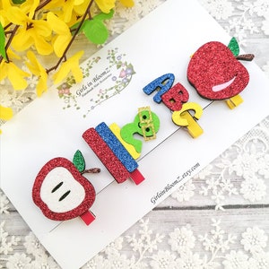 May include: Set of five glitter hair clips featuring a red apple, a stack of books, a green worm with glasses, and the letters A, B, and C. The clips are on a white background with a small business logo.