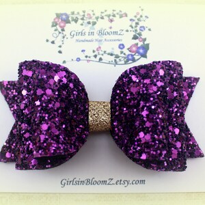 May include: Purple and gold glitter hair bow. The bow is made of two layers of purple glitter fabric with a gold glitter center. The bow is attached to a clip.