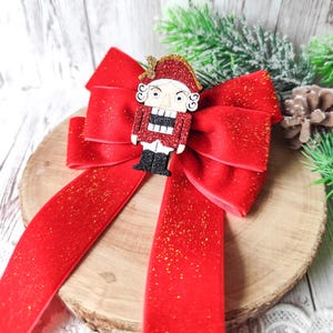 Op de afbeelding: Een feestelijke rode fluwelen strik met gouden glitteraccenten, versierd met een glitterende notenkrakerfiguur. De strik ligt op een houten schijf en dennentakken, perfect voor de kerstversiering.