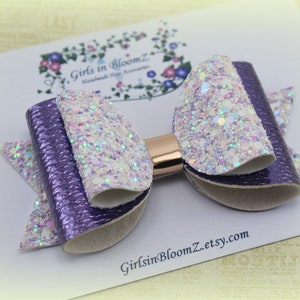 May include: A handmade hair bow featuring a layered design with purple and glittery white accents. The bow has a metallic gold center and is set against a white background with the text "Girls in BloomZ etsy.com".