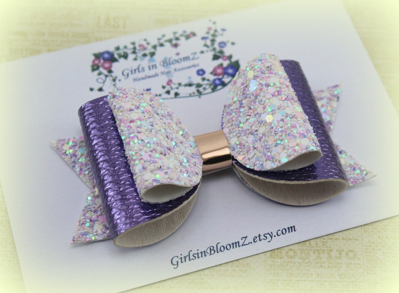 May include: A handmade hair bow featuring a layered design with purple and glittery white accents. The bow has a metallic gold center and is set against a white background with the text "Girls in BloomZ etsy.com".