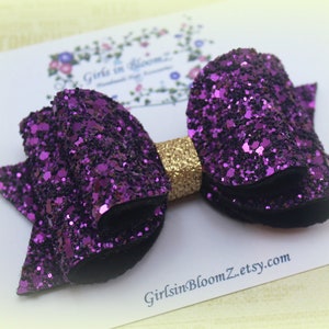 May include: A purple glitter hair bow with a gold center. The bow is on a white background with the text "GirlsInBloomZ.etsy.com".