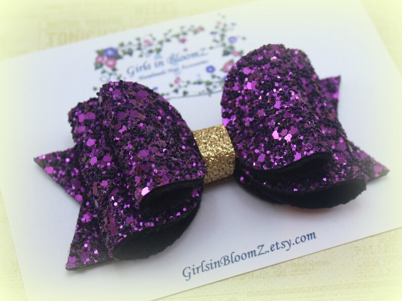 May include: A purple glitter hair bow with a gold center. The bow is on a white background with the text "GirlsInBloomZ.etsy.com".